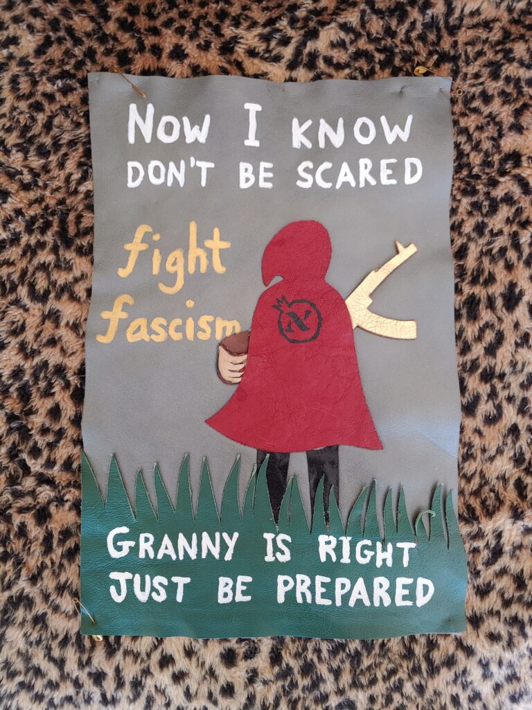 leather applique of little red riding hood, from behind, holding a basket and a gold AK-47 & with a circle-alef on her cape, with "now i know, don't be scared / granny is right, just be prepared" and "fight fascism" written around her